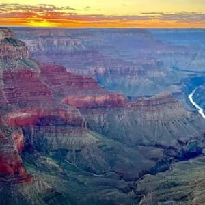 A wide canyon with layered red rock formations and a winding river seen at sunset under an orange sky.