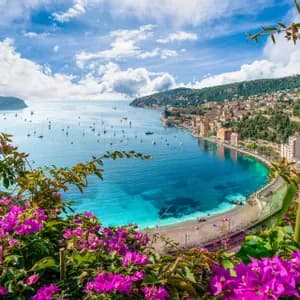Una vista dall'alto di una cittadina costiera lungo una baia turchese punteggiata di barche, incorniciata da vivaci fiori rosa sotto un cielo azzurro nuvoloso.