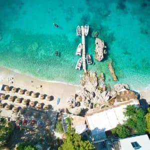 Luftaufnahme eines sonnigen Strandes mit klarem türkisen Wasser, einem Holzsteg mit Booten und Reihen von Strohschirmen.