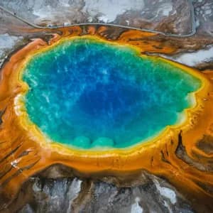 An aerial, top-down view of a large geothermal hot spring with multicolored rings of orange and yellow around blue water.