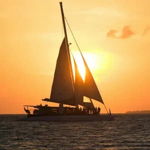 A WeRoad group trip on a catamaran is silhouetted on the ocean against a vibrant orange sunset.