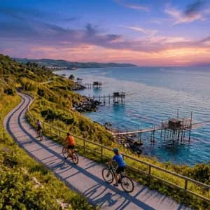 Veduta aerea di un viaggio di gruppo WeRoad in bicicletta su un sentiero costiero tortuoso con tradizionali moli da pesca sul mare al tramonto.