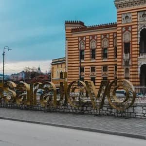 Una grande insegna dorata 'Sarajevo' si erge su un marciapiede davanti a un edificio storico a strisce sotto un cielo nuvoloso.