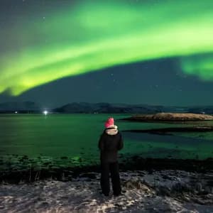 Una persona con un gorro rosa de pie en una orilla nevada observando la aurora boreal verde iluminar el cielo nocturno sobre una gran masa de agua.