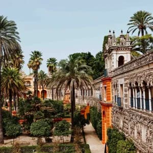 An ornate stone building with a long, arched balcony overlooks a lush garden filled with tall palm trees and pathways.