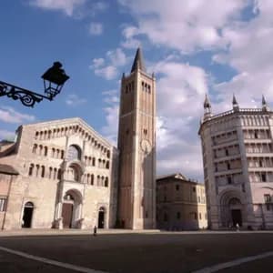 Una storica cattedrale in pietra, campanile e battistero in una grande piazza assolata sotto un cielo blu con nuvole.