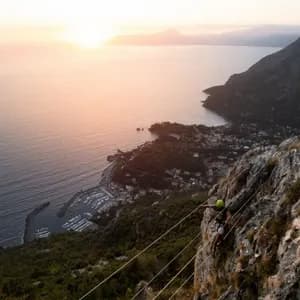 Una persona che scala una via ferrata su una scogliera, con una vista aerea di un porto turistico e della costa al tramonto.