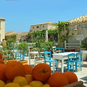 Una cassa di arance fresche in primo piano in un caffè all'aperto soleggiato con sedie blu vivaci e tavoli bianchi.