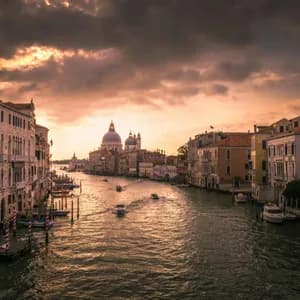 Un ampio canale con barche è fiancheggiato da edifici storici sotto un cielo drammatico al tramonto, con una basilica a cupola in lontananza.