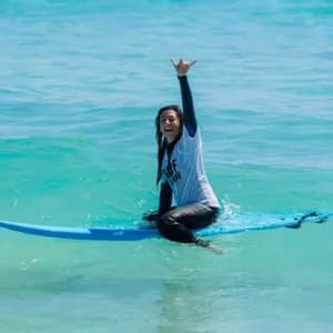 Una donna sorridente in muta siede su una tavola da surf blu in acqua turchese, facendo il segno shaka con la mano.