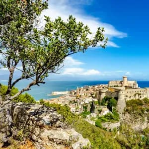 Una storica fortezza e città su una scogliera rocciosa a picco sul mare blu, vista da un punto panoramico dietro un albero.