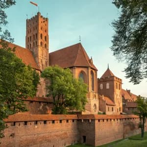 Un grande castello medievale in mattoni rossi con un'alta torre e mura fortificate, circondato da alberi verdi al tramonto.