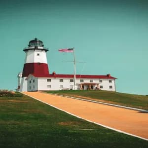 A red and white lighthouse with an adjacent building sits atop a grassy hill under a clear teal sky, with a path leading up to it.