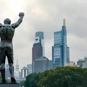 The back of a bronze boxer statue with arms raised in victory, overlooking a modern city skyline under an overcast sky.