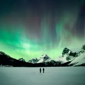 Two people stand on a vast snowy landscape watching the green and purple aurora borealis over snow-capped mountains.