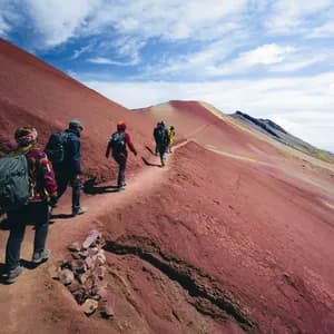 Eine WeRoad-Gruppe wandert im Gänsemarsch auf einem schmalen Pfad am Hang eines riesigen, roten Berges unter teils bewölktem Himmel.