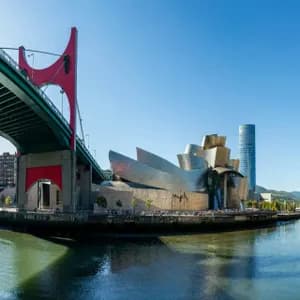 A panoramic view of a modern metallic museum and a red bridge over a river on a sunny day.