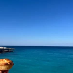 Una mano tiene un piccolo pasticcino davanti a un mare turchese con lontani edifici costieri bianchi sotto un cielo azzurro limpido.