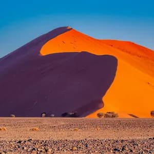A large orange sand dune casts a deep shadow across itself in a rocky desert landscape under a clear blue sky.