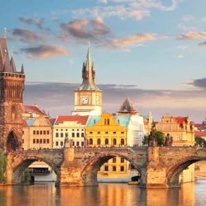 Un pont en arc en pierre historique avec une tour gothique enjambe une rivière devant la ligne d'horizon d'une ville européenne au coucher du soleil.