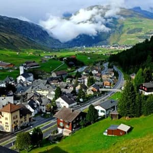 Una vista aerea di un villaggio incastonato in una lussureggiante valle di montagna verde, con una strada che lo attraversa sotto un cielo parzialmente nuvoloso.