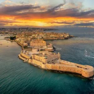 An aerial view of a stone fortress on a peninsula and a coastal city during a colorful sunset over the sea.