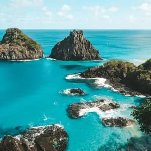 Due grandi formazioni rocciose, ricoperte di verde, si ergono da un oceano turchese vicino a una costa rocciosa sotto un cielo parzialmente nuvoloso.