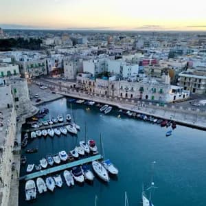Una vista aerea di una città costiera al tramonto, con molte barche ormeggiate in un piccolo porto accanto a una storica torre di pietra.