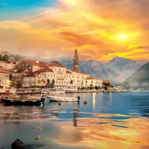 Una storica cittadina costiera con un campanile si affaccia su un porto sereno con barche, incorniciata dalle montagne sotto un tramonto dorato.
