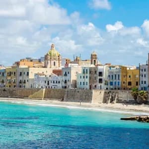 Una città costiera con edifici colorati e una muraglia in pietra si affaccia su una spiaggia sabbiosa con acqua turchese sotto un cielo parzialmente nuvoloso.