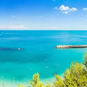 Vista aerea di una spiaggia sabbiosa affollata con ombrelloni colorati e persone che nuotano in acqua turchese in una giornata di sole.