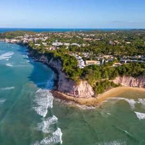 Vista aérea de un pueblo costero sobre acantilados rojizos, con vegetación exuberante, una playa de arena y el océano turquesa debajo.