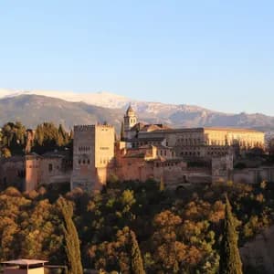 Una grande fortezza in pietra su un fianco di collina boscoso, illuminata da una luce dorata, con montagne innevate sullo sfondo.