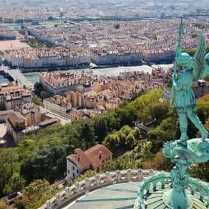 Una statua verde di un angelo alato su una cupola si affaccia su un paesaggio urbano denso con un fiume e edifici dai tetti in terracotta.