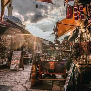 Un callejón empedrado bañado por el sol está bordeado de puestos de mercado que venden artesanías tradicionales, con una cúpula visible contra un atardecer nublado.