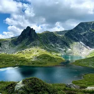 Un lac alpin aux eaux d'un bleu profond est niché dans une vallée entourée de collines verdoyantes et de sommets rocheux sous un ciel nuageux.