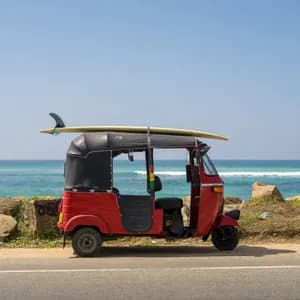 Un tuk-tuk rosso con una tavola da surf legata al tetto è parcheggiato a bordo strada vicino all'oceano sotto un cielo azzurro e limpido.