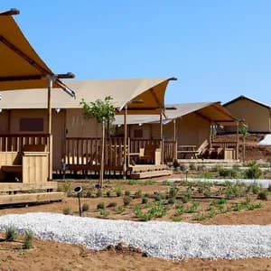 Una fila di tende glamping con verande in legno si trova lungo un sentiero di ghiaia bianca in un'area paesaggistica sotto un cielo azzurro e limpido.