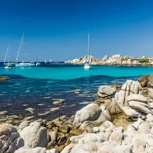 Un littoral rocheux surplombe une baie calme avec des voiliers sur une eau turquoise claire sous un ciel bleu éclatant.