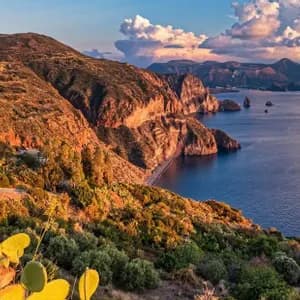 Una vista panoramica di una costa frastagliata con fichi d'India, illuminata dalla luce dorata del tramonto sul mare.