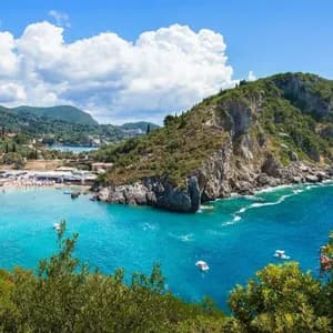 Una vista elevada de una playa concurrida con agua turquesa en una cala, rodeada de acantilados cubiertos de árboles y montañas.