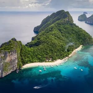 Aerial view of a lush, rocky island featuring a white sand beach with many boats moored in the surrounding turquoise water.