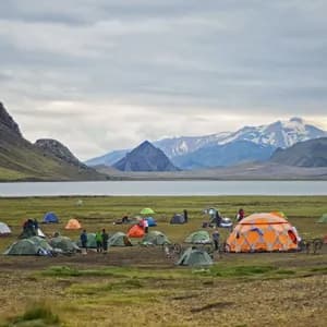Un voyage de groupe WeRoad avec des tentes colorées et des vélos, campant au bord d'un lac au pied de montagnes escarpées et enneigées.