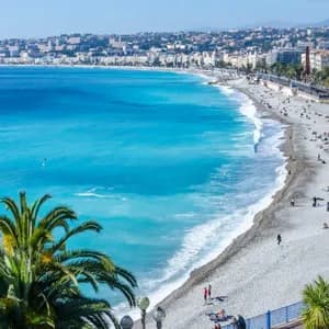 A panoramic view of a coastal city with a long pebble beach, turquoise water, and a promenade lined with palm trees.