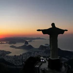 The silhouette of a large statue with outstretched arms overlooks a coastal city, a bay, and mountains during a colorful sunrise.