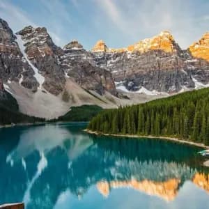 Montagne innevate illuminate dal sole si riflettono in un lago turchese calmo circondato da una fitta foresta di pini.