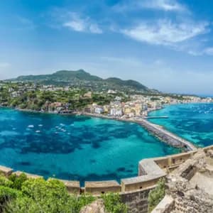 A high-angle, panoramic view of a coastal town and causeway surrounded by turquoise sea, seen from a stone fortification.