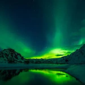 L'aurora boreale verde brilla in un cielo stellato sopra montagne innevate, il suo riflesso visibile nelle calme acque sottostanti.