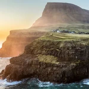 Una cascata si getta da una verde scogliera erbosa nell'oceano, con un villaggio sullo sfondo al tramonto.