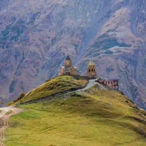 Una storica chiesa in pietra e un campanile appollaiati su una collina erbosa di fronte a una vasta catena montuosa rocciosa.